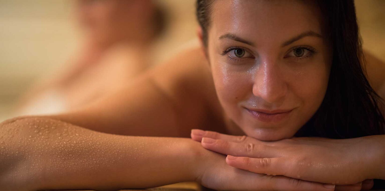Smiling woman resting her chin on her hands in a warm, softly lit sauna