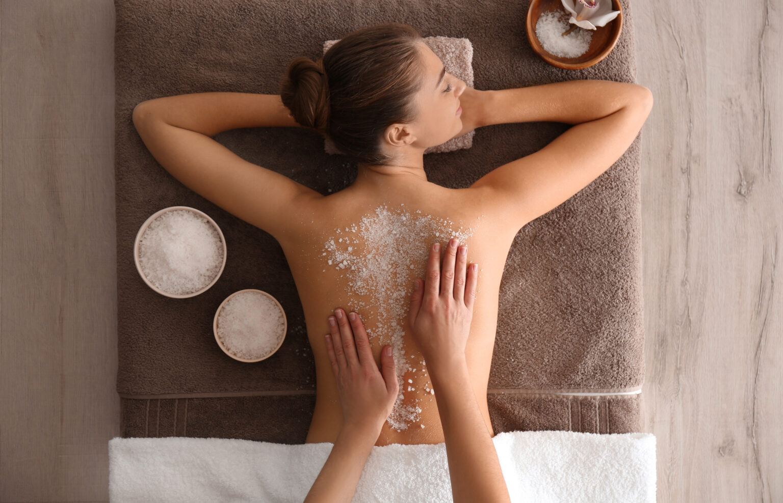 Woman lying face down on a spa table while a therapist applies a salt scrub to her back