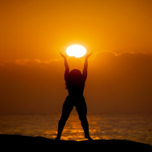 Silhouette of a person raising both arms toward a glowing sun at sunset over the water